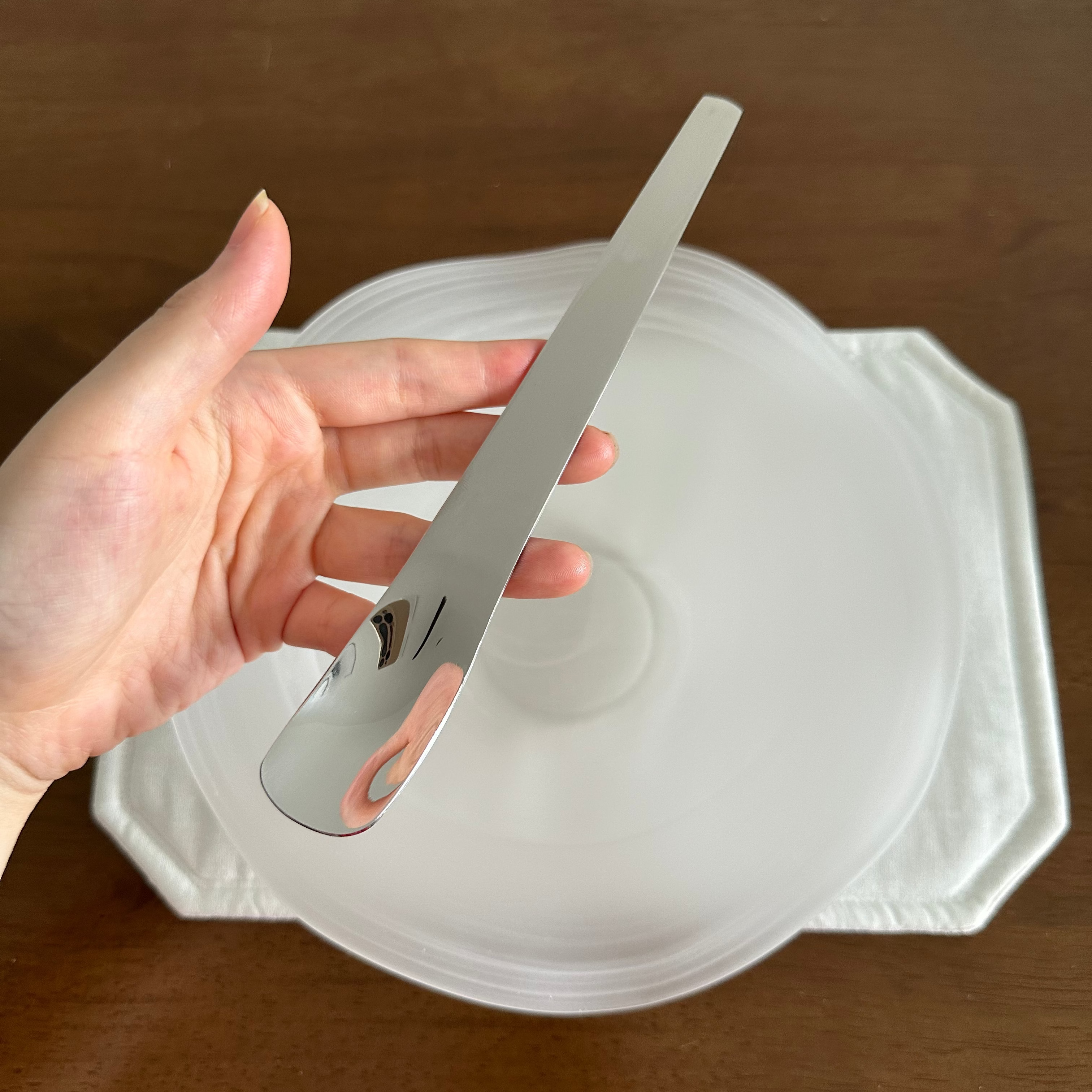 Hand holding a silver spoon over a white plate on a wooden surface