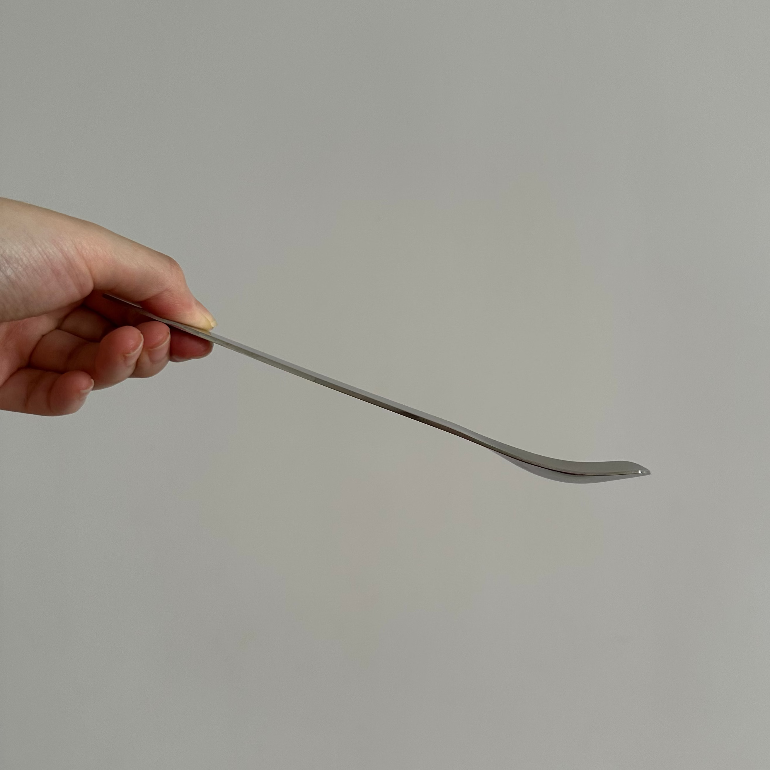 Hand holding a long, thin stainless steel spoon against a plain background