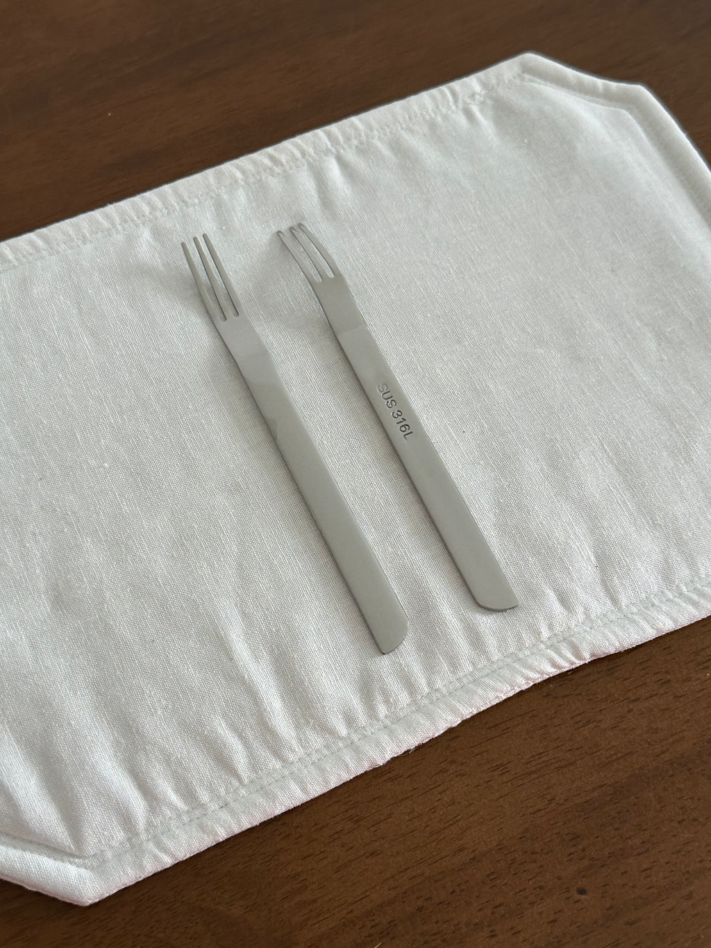 Two silver forks on a white cloth with sus 316L on a wooden surface.